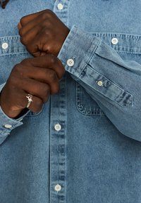 Lichtblauwe denimshirts met een voorzak, knoopsluiting en witte knopen; de stof heeft een gladde textuur.