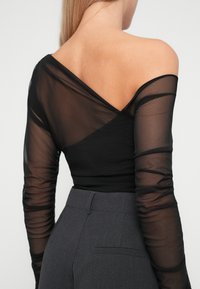 Top lungo a maniche nere in rete con design off-the-shoulder, caratterizzato da maniche trasparenti e corpetto aderente, abbinato a pantaloni grigi a vita alta.