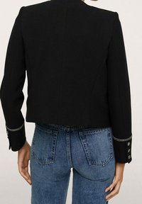 Czarna krótka kurtka z wysokim kołnierzem i ozdobnymi guzikami na mankietach, zestawiona z niebieskimi dżinsami z tylnymi kieszeniami. Gładka faktura.