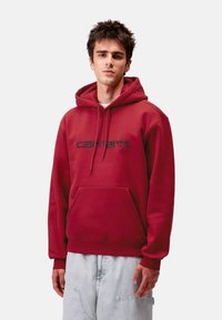 Sudadera de algodón roja con un bolsillo tipo canguro en la parte frontal y cordones. Presenta un logo azul marino en el pecho. Textura suave, corte relajado.