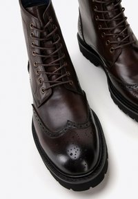 Hnědé kožené kotníkové boty s tkaničkami, designem wingtip, brogue detaily a silnou černou gumovou podrážkou. Hladký povrch s lesklým finišem.