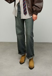 Veste en cuir marron, chemise rayée, cravate bleu marine et rouge, jean en denim délavé avec ourlets retroussés, et bottines jaunes à bouts pointus.