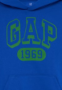 Felpa blu con grande scritta "GAP" verde e blu e "1969" all'interno di un ovale verde arrotondato. Il materiale sembra morbido con una vestibilità standard.
