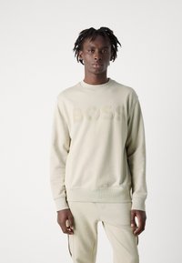 Sudadera crema con un logo texturizado de "BOSS", cuello redondo, puños de canalé y un corte relajado. Se combina con pantalones crema a juego.
