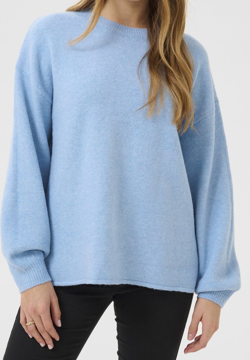 Femme portant un pull bleu clair oversize avec de longues manches bouffantes et un pantalon noir, debout devant un fond uni.