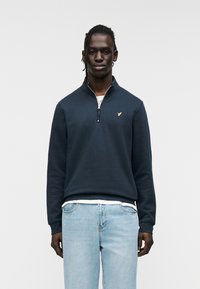 Μπλε ναυτικό φούτερ με γιακά μισής ζώνης, με διακριτικό χρυσό λογότυπο στο στήθος. Ριμπ μανσέτες και τελείωμα. Ανοιχτό μπλε τζιν.