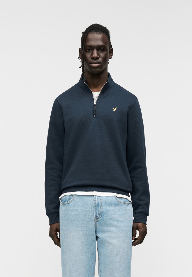 Μπλε ναυτικό φούτερ με γιακά μισής ζώνης, με διακριτικό χρυσό λογότυπο στο στήθος. Ριμπ μανσέτες και τελείωμα. Ανοιχτό μπλε τζιν.