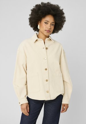 Femme avec une coiffure afro naturelle portant une veste crème boutonnée et un jean bleu foncé, debout devant un fond gris clair uni.