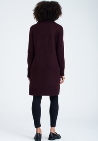 Burgundy stickad klänning med hög krage, ribbade ärmslut och fåll. Bärs över svarta tajta byxor och svarta skor.