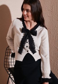 Ivory cardigan with black lace trim and bow, featuring six buttons and flared cuff details. Designed with a V-neck and high-low hem.