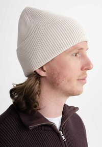 Witte geribbelde gebreide beanie met een puntige bovenkant, gedragen door een persoon met schouderlang haar. Achtergrond is vlak wit.