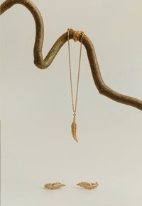Collier en forme de feuille en or avec un pendentif texturé, accompagné de clous d'oreilles assortis en forme de feuille. Exposé sur une branche courbée sur un fond clair.
