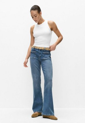 Vrouw die een witte mouwloze top draagt, blauwe high-waisted uitlopende spijkerbroek met voorzakken, een lichtbruine riem en bruine schoenen, staand tegen een effen achtergrond.