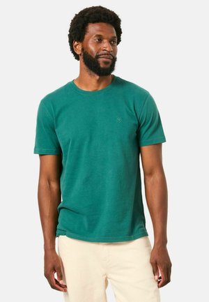 Mężczyzna z brodą, ubrany w morski t-shirt z krótkim rękawem i jasnobeżowe spodnie, stojący i lekko patrzący na bok na jednolitym tle.