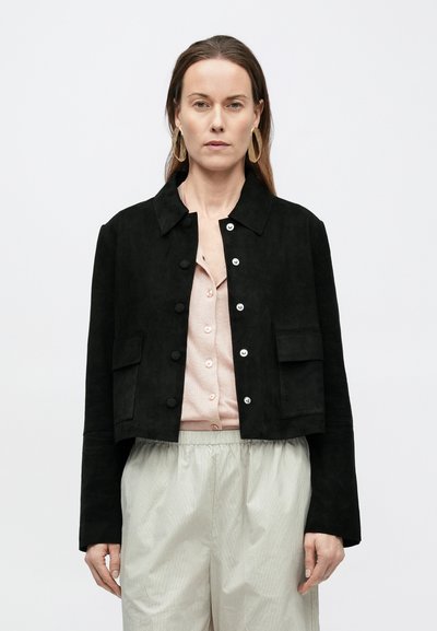 Μαύρο τζάκετ cropped από σουέτ με κουμπιά μπροστά, δύο μπροστινές τσέπες και μυτερό γιακά, φορεμένο πάνω από ανοιχτό ροζ μπλουζάκι με κουμπιά.