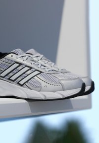 Biele atletické topánky s panelmi z sieťoviny, čiernymi akcentmi a tromi čiernymi pruhmi. Majú gumovú podrážku a hladký textúrovaný zvršok.