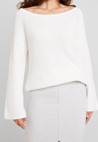 Žena v bílém svetr s odhalenými rameny a ve světle šedé úzké sukni na jednobarevném pozadí.