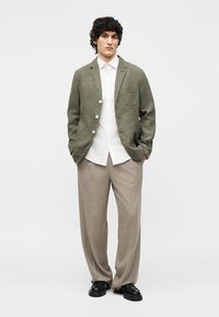 Άνδρας με σγουρά μαλλιά που φοράει σακάκι χακί, λευκό πουκάμισο, μπεζ παντελόνι με φαρδιά μπατζάκια και μαύρα παπούτσια, στέκεται με τα χέρια στις τσέπες.