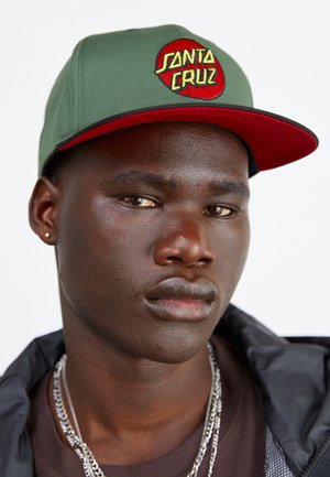 Jeune homme portant une casquette Santa Cruz verte et rouge, une veste sombre, une chemise marron et des chaînes argentées, avec une petite boucle d'oreille à l'oreille gauche.