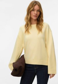Pull jaune en maille avec un ourlet côtel é et des manches larges, accompagné d'un sac à main en tissu marron. Porté avec un jean bleu foncé.