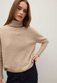 Beige Rollkragenpullover aus weichem Strickmaterial, mit einer lockeren Passform, gerippten Bündchen und einem seitlichen Schlitz am Saum.