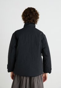 Schwarze Daunenjacke mit hohem Kragen, versehen mit einem strukturierten Stoff und einer lockeren Passform. Die Jacke hat lange Ärmel und schräge Nähte.