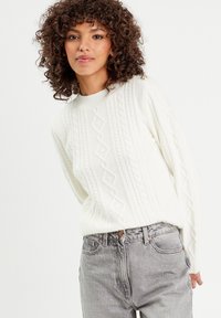 Pull en maille blanche avec des motifs en diamant texturés, associé à un jean en denim gris clair. La texture est douce et épaisse.