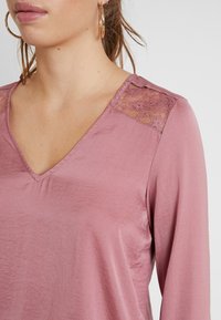 Pitkähihainen V-pääkaita pehmeässä, vaaleanpunaisessa kankaassa, jossa pitsikoristus hartioilla, sileä pinta ja rento malli.