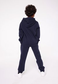 Felpa blu navy con cappuccio e pantaloni della tuta coordinati, realizzati in tessuto morbido, con polsini a coste e vestibilità rilassata, abbinati a sneakers bianche.