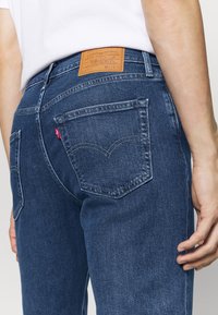 Blå denimjeans med rak skärning, med en brun läderflik, två bakfickor med sömmar och en liten röd logotyp på vänster ficka.