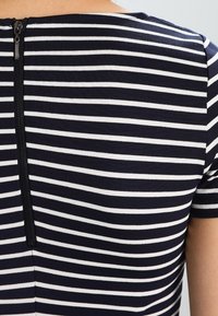 T-shirt rayé bleu marine et blanc à manches courtes, avec une fermeture éclair dans le dos et un col rond. Texture de tissu lisse.