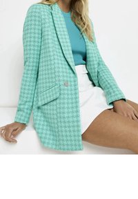 Blazer strutturato teal chiaro con motivo a pied-de-poule, dettaglio di tasca singola e accentuazione hardware decorativa. Indossato sopra un top blu e una gonna bianca.