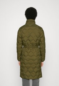 Personne portant un long manteau matelassé vert olive avec un col haut, une taille élastique et des manches longues, de dos contre un fond uni.