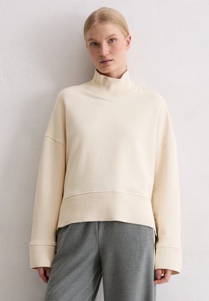 Cream oversized sweatshirt with high neck, ribbed cuffs, and a textured waistband, paired with gray pants. Soft fabric, minimal design.