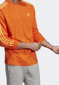 Camiseta de algodón naranja de manga larga con tres franjas blancas en cada hombro y un peque ño logotipo en el pecho.