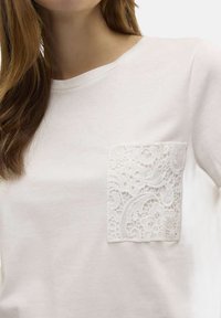 T-shirt en coton blanc avec une poche en dentelle texturée sur le côté gauche, un col rond et des manches courtes.