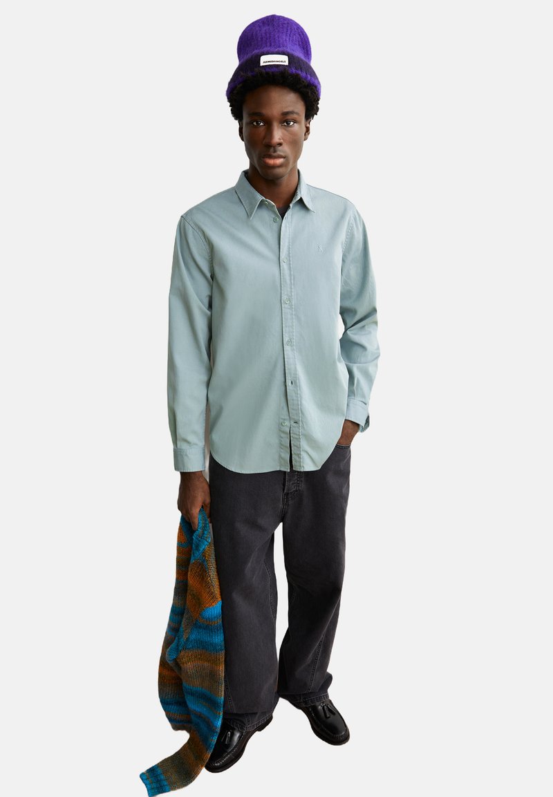 Hellgrüne Button-Up-Hemd, dunkelgraue lockere Hose, mit einer lila Strickmütze, hält einen mehrfarbigen gestreiften Pullover. Schwarze Schuhe.
