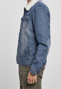 Hombre que lleva una chaqueta de mezclilla azul con cuello de forro polar blanco y pantalones cargo verdes oliva, de pie de lado con el brazo relajado.