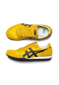 Žluté semišové tenisky s černými pruhy, mají ploché šňůrky, texturovanou podrážku a štítek s logem na jazyku. Neformální design a lehká konstrukce.