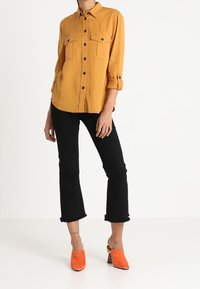 Senapsgul knäppt skjorta med två bröstfickor, svarta utsvängda jeans med fransad fåll, och orange klackskor.