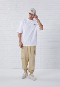 White oversized t-shirt with black printed text, beige cargo pants with side pockets, and light sneakers; model wearing a light gray cap.