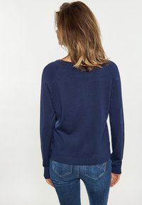 Dunkelblauer Strickpullover mit lockerer Passform, langen Ärmeln und gerippten Bündchen, kombiniert mit schmalen Jeans, die ein dezentes Design der hinteren Taschen aufweisen.