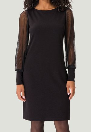 Femme portant une robe noire longueur genoux avec des manches longues en voile transparent et des poignets opaques, debout devant un fond clair uni.