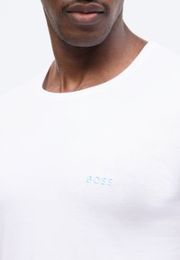 Nærbillede af en mands nedre ansigt og nakke iført en hvid T-shirt med lyseblå "BOSS"-tekst på brystet.