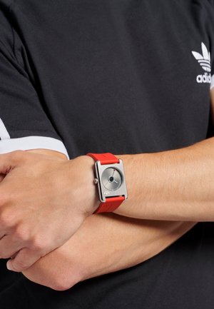 Square-shaped watch with a silver metal case, dark dial, and red silicone strap, featuring a minimalist design and a single subdial.