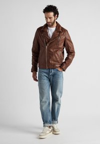 Brun lädermotorcykeljacka med spetsig krage, dragkedja fram och sidofickor, tillsammans med ljusblå denim och krämfärgade sneakers.