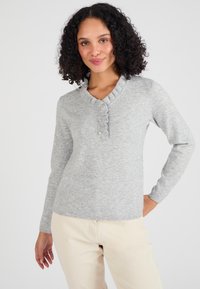 Maglione grigio a maglia con colletto arricciato, quattro bottoni sul davanti e maniche lunghe. Abbinato a pantaloni chiari.
