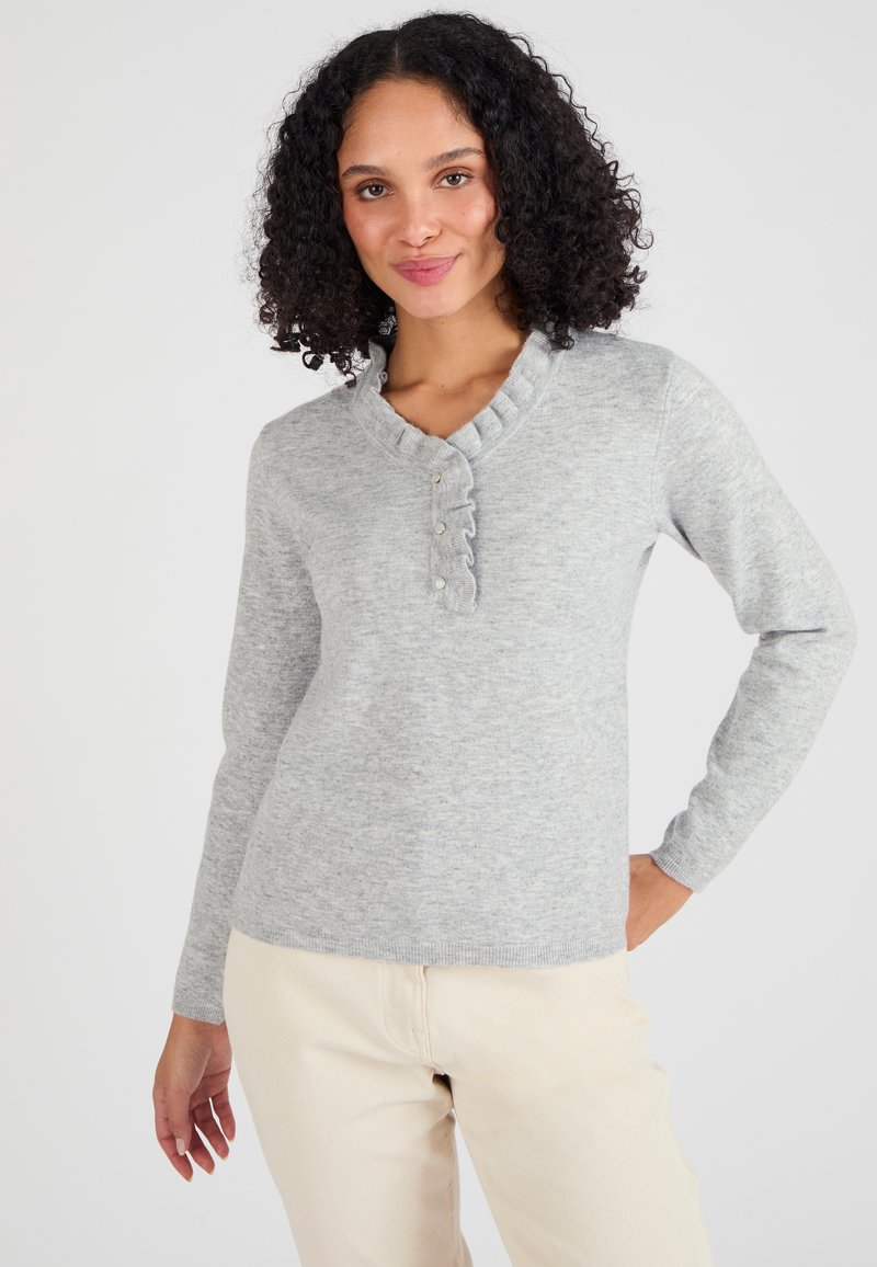 Suéter de punto gris con cuello volante, cuatro botones en el frente y mangas largas. Combinado con pantalones de color claro.