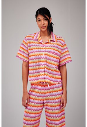 Vrouw draagt een blouse met kraag en bijpassende broek met een roze, oranje en witte zigzagpatroon, staand tegen een effen achtergrond.