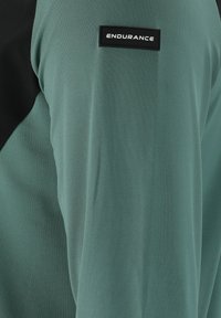 Grünes Performance-Shirt mit einem schwarzen Schulterbereich und einem kleinen schwarzen Etikett mit dem Text "ENDURANCE." Weiche, glatte Textur.